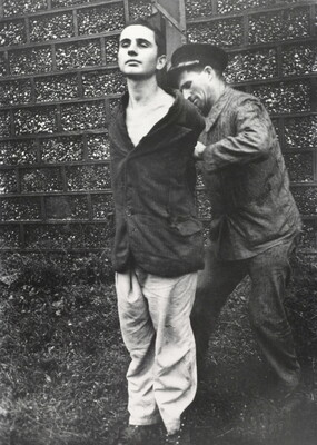 A black-and-white photograph of a young, White man standing with his hands behind his back as another White man ties them to a wood pole.