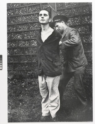 A black-and-white photograph of a young, White man standing with his hands behind his back as another White man ties them to a wood pole.