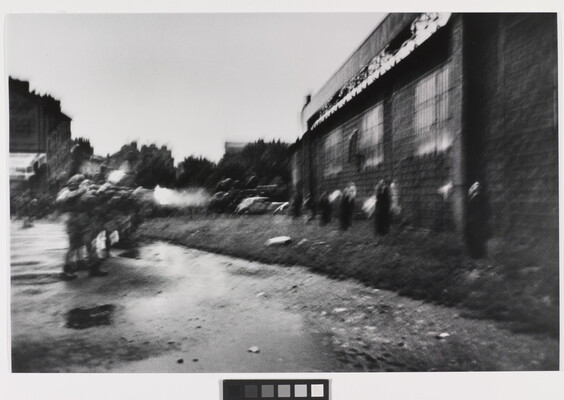 A blurry black-and-white photograph of soldiers on the left shooting rifles at a line of people standing against a wall on the right.