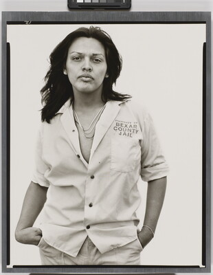 A black-and-white photograph of a medium-skinned person, with long hair wearing a light, partially unbuttoned shirt with text "Property of Bexar County Jail" above the breast pocket.