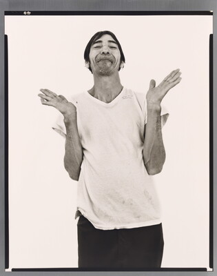 A black-and-white portrait photograph of a medium-skinned man with short hair, light facial hair, wearing a white t-shirt, dark pants, and hands open palmed and raised to shoulders.
