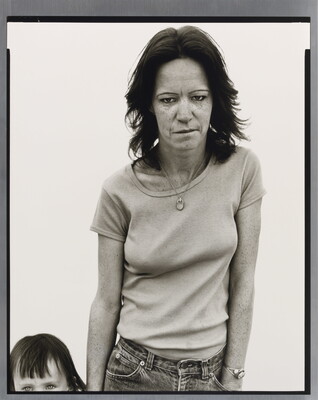 A black-and-white portrait photograph of a weary-looking White woman standing beside a child whose head and body is mostly out of frame.