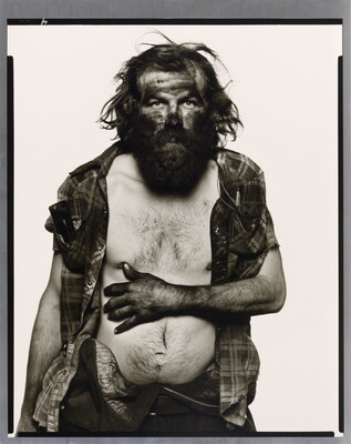 A black-and-white portrait photograph of a bearded man, head and arms covered in dark coal dust, resting his arm across his bare white chest.