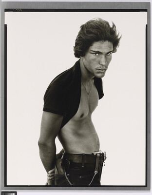 A black-and-white portrait photograph of a medium-skinned man standing sideways, posture in a subtle S-curve, arms behind his back, with a dark t-shirt pulled up behind his neck.