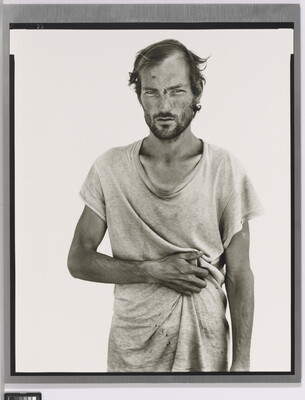 A black-and-white portrait photograph of a White man wearing a loose-fitting dirty white t-shirt, resting his arm across his stomach.