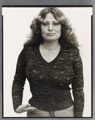 A black-and-white portrait photograph of a White woman with long hair curled back from her face, wearing a fitted dark sweater, and one hand in her pants pocket.