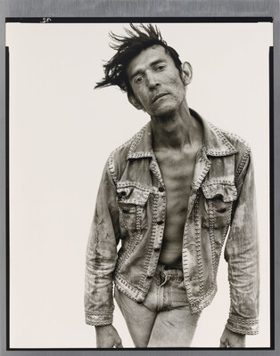 A black-and-white portrait photograph of a White man with dark hair that sticks up, wearing jeans and a jean jacket, and crossing his legs while bending forward.