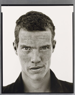 A black-and-white close-up photograph of a White man with short cropped hair, freckles across his face, wearing a dark collared shirt.