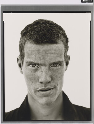A black-and-white close-up photograph of a White man with short cropped hair, freckles across his face, wearing a dark collared shirt.