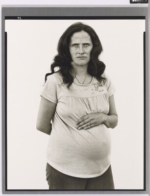 A black-and-white portrait photograph of a pregnant White woman with long, dark hair placing one arm across her stomach and the other behind her back.