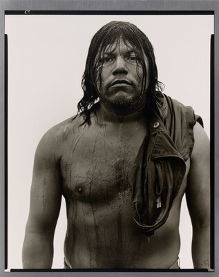 A black-and-white portrait photograph of a stocky, dark-skinned man with long, dark wet hair that drips down his shirtless torso.