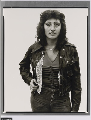 A black-and-white portrait photograph of a medium-skinned woman with dark, feathered hair wearing jeans, a jacket, V-neck top, and a heart necklace.