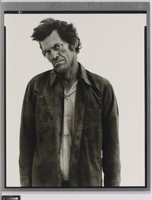 A black-and-white portrait photograph of a White man standing with his head cocked, frowning, and wearing two soiled shirts.