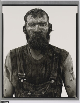 A black-and-white portrait photograph of a bearded man covered in mud and dirt, wearing dirty overalls, and looking straight at the camera.