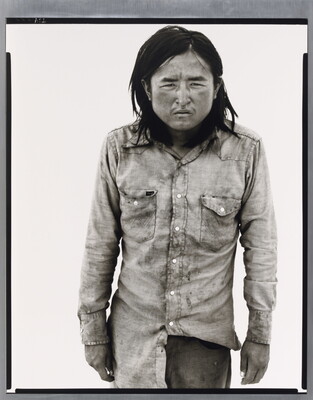 A black-and-white portrait photograph of a medium-skinned man with long hair, wearing dirty clothes, and squinting straight at the camera.