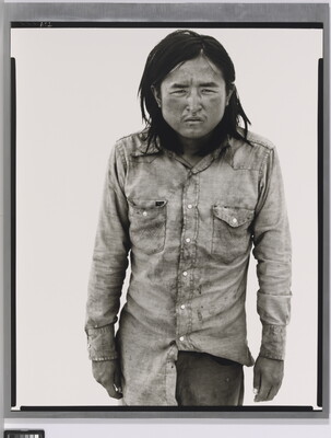 A black-and-white portrait photograph of a medium-skinned man with long hair, wearing dirty clothes, and squinting straight at the camera.