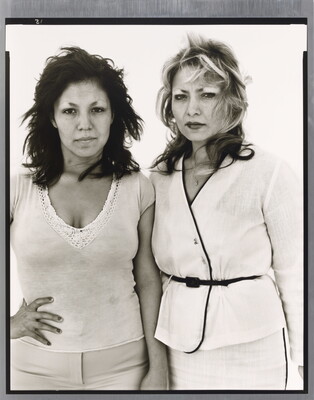 A black-and-white portrait photograph of two medium-skinned women, one in white pants and top with a hand on her hip and the other in a belted white suit, both looking intensely at the camera.