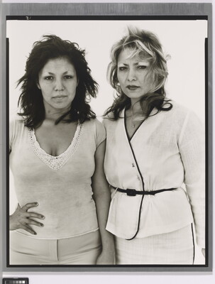 A black-and-white portrait photograph of two medium-skinned women, one in white pants and top with a hand on her hip and the other in a belted white suit, both looking intensely at the camera.