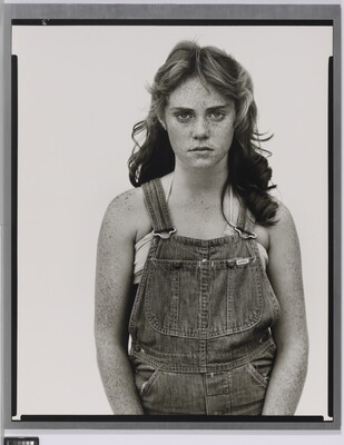 A black-and-white portrait photograph of a young White girl with wavy hair, freckles on her face and arms, wearing denim overalls.