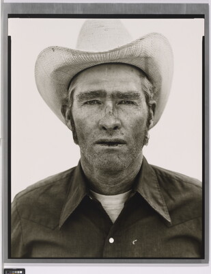 A black-and-white portrait photograph of a White man wearing a cowboy hat and collared shirt, with stubble on his face and chapped lips.