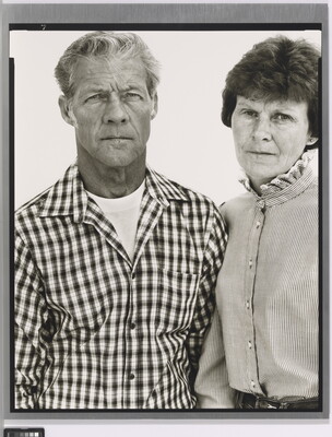 A black-and-white portrait photograph of a middle-aged White man wearing a checked button-down shirt and a middle-aged White woman in a striped high-collar blouse standing shoulder to shoulder.