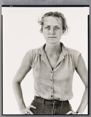 A black-and-white portrait photograph of a young White woman in a sleeveless blouse, hair pulled back from her face, and hands on her hips.