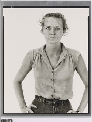 A black-and-white portrait photograph of a young White woman in a sleeveless blouse, hair pulled back from her face, and hands on her hips.