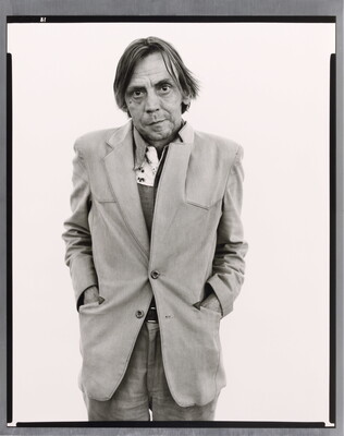 A black-and-white portrait photograph of a middle-aged White man with his hands in the pockets of his unkempt suit jacket.