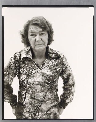A black-and-white portrait photograph of an older White woman with wavy hair wearing a patterned polo shirt, hands hooked into her back pants pockets.