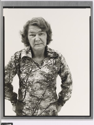 A black-and-white portrait photograph of an older White woman with wavy hair wearing a patterned polo shirt, hands hooked into her back pants pockets.