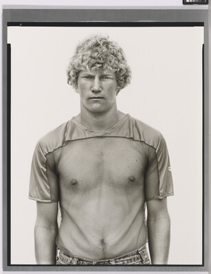 A black-and-white photograph of a young White man with curly blonde hair wearing a see-through, mesh shirt.