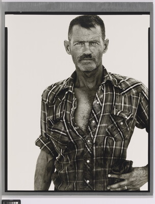 A black-and-white portrait photograph of a middle-aged White man with scruffy facial hair wearing a plaid button-down shirt standing with one hand on his hip.