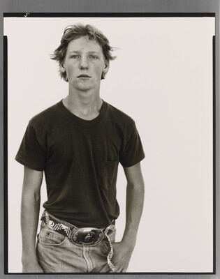 A black-and-white portrait photograph of a White teenage boy in a dark t-shirt and belted jeans with a large belt buckle, one hand hooked into his jeans pocket.