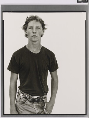 A black-and-white portrait photograph of a White teenage boy in a dark t-shirt and belted jeans with a large belt buckle, one hand hooked into his jeans pocket.