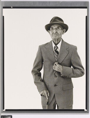 A black-and-white portrait photograph of an elderly White man in a hat, suit, tie, and one hand holding a cane.