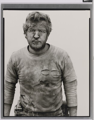 A black-and-white portrait photograph of a White man with messy hair and a beard wearing a dirty, ripped shirt.