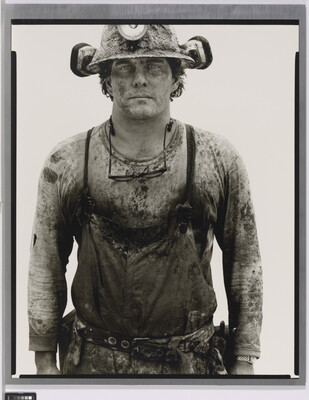 A black-and-white portrait photograph of a young White man covered in dirt wearing a hard hat and a shirt under coveralls.