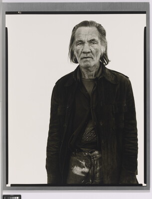 A black-and-white portrait photograph of an older White man with long hair wearing a partially-buttoned jacket over his clothing.