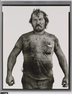 A black-and-white portrait photograph of a White man with messy hair and a beard wearing a dirty t-shirt and holding his arms out from his sides.