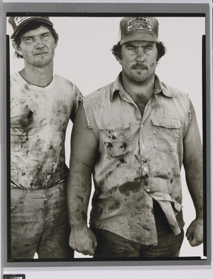 A black-and-white portrait photograph of two White men wearing dirty clothes and baseball caps standing next to each other.