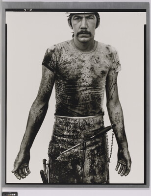 A black-and-white portrait photograph of a White man with a mustache wearing a dirty t-shirt and apron holding with different tools on it.