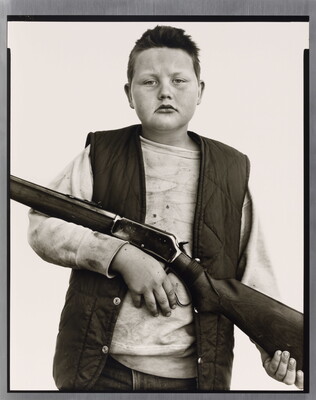 A black-and-white portrait photograph of a White boy with close cropped hair, wearing a quilted vest over a sweatshirt, and holding a rifle across his torso.