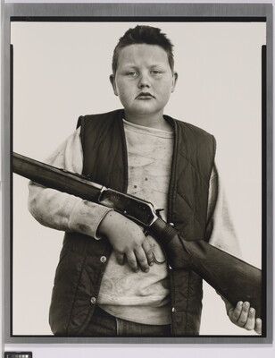 A black-and-white portrait photograph of a White boy with close cropped hair, wearing a quilted vest over a sweatshirt, and holding a rifle across his torso.