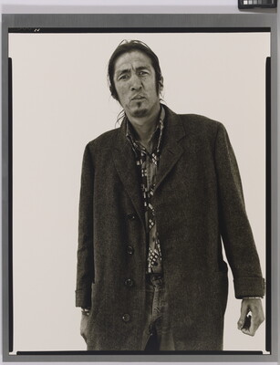 A black-and-white portrait photograph of a medium-skinned man with long hair, facial hear wearing a long coat.