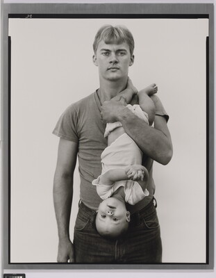A black-and-white portrait photograph of a White man with light hair holding a baby upside down in front of his chest.