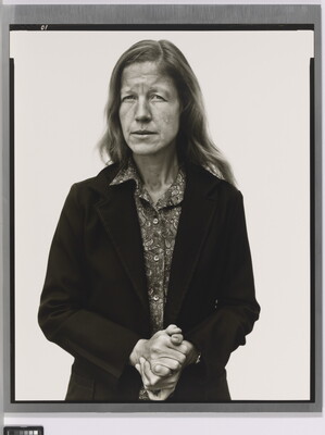 A black-and-white portrait photograph of a White woman with long hair wearing a button-down blouse under a suit jacket holding her hands clasped in front of her.
