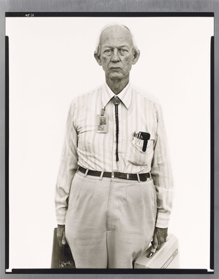A black-and-white portrait photograph of an older, mostly bald White man wearing a collared shirt, a bolo tie, and work badge attached to his collar, holding two briefcases.