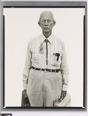 A black-and-white portrait photograph of an older, mostly bald White man wearing a collared shirt, a bolo tie, and work badge attached to his collar, holding two briefcases.