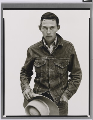 A black-and-white portrait photograph of a young White man with short dark hair wearing a denim jacket and holding a cowboy hat in front of him.