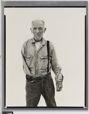 A black-and-white portrait photograph of an older, balding White man with one arm wearing a button-down shirt and jeans held up with one suspender.